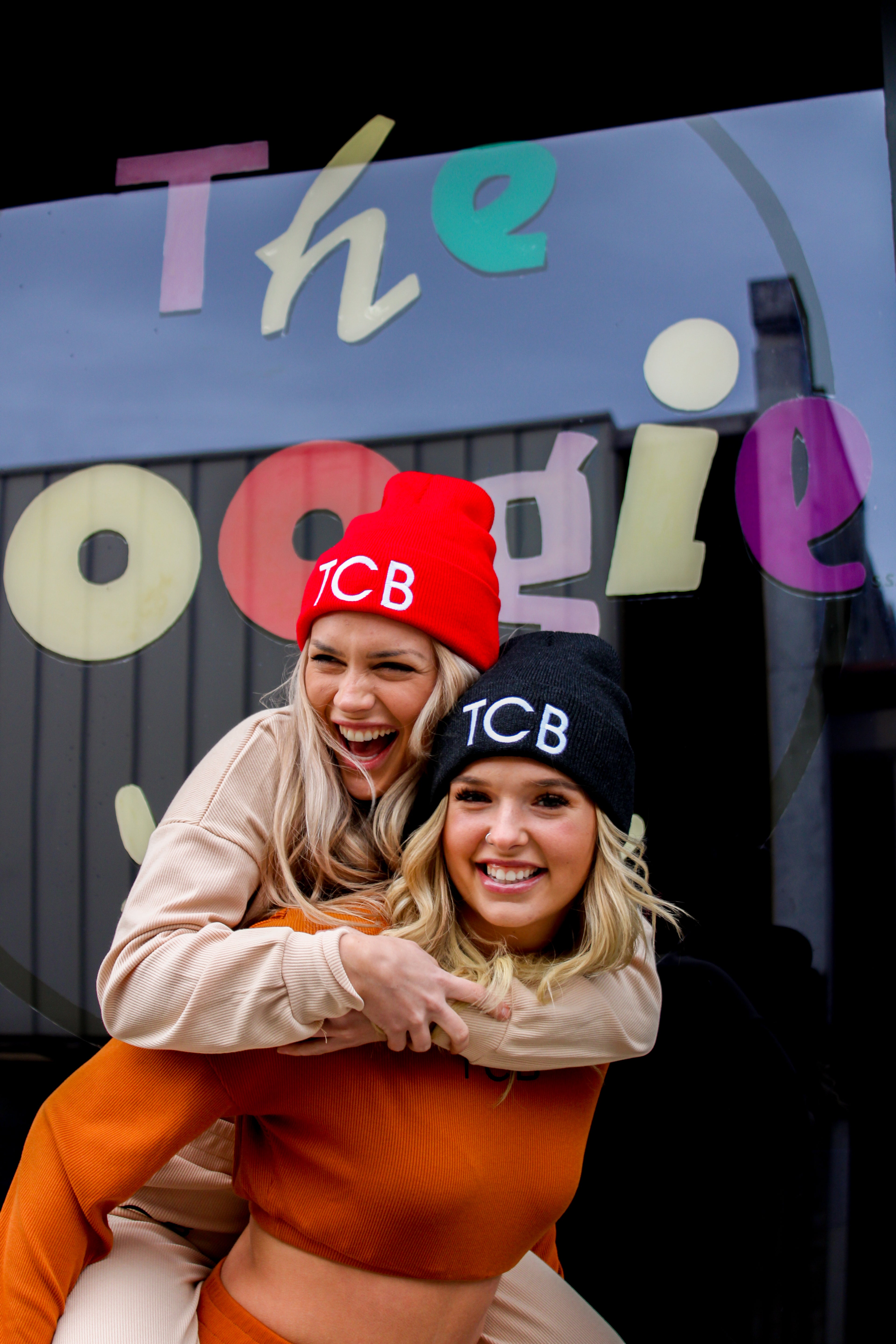 Two people wearing red and black beanies with 'TCB' on them, standing in front of a colorful sign.